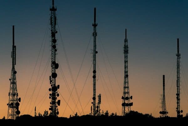 Silhouetted communication towers stand against a vibrant sunset sky. The gradient transitions from orange to deep blue, creating a serene mood. cybersecurity