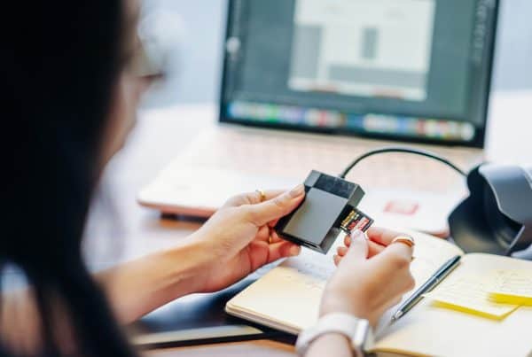 A person inserts a memory card into a reader next to a laptop with blurred content on the screen. An open notebook and pen are on the desk. managed file transfer