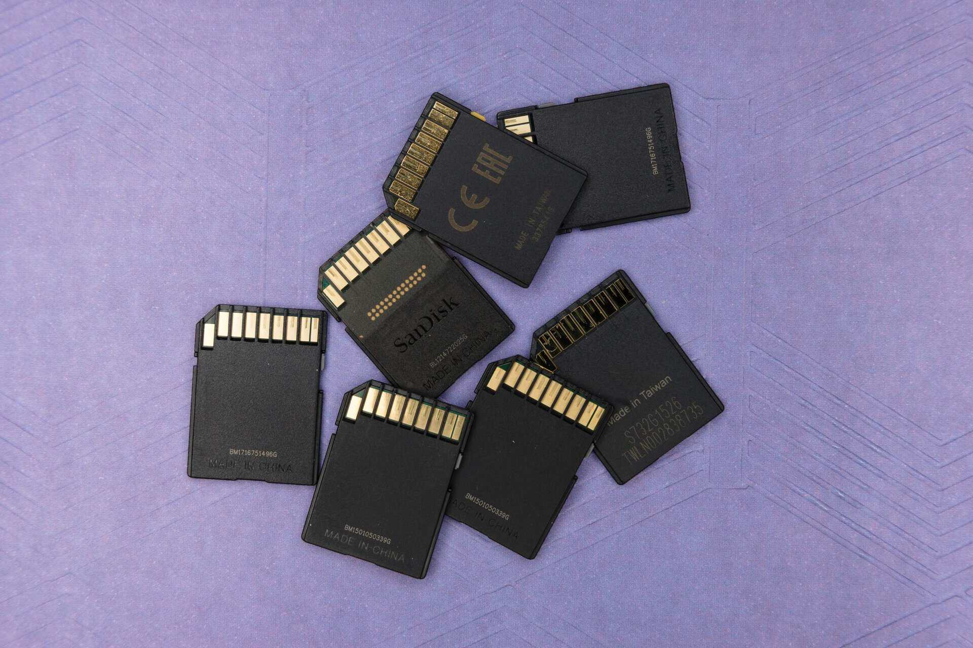 Seven black SD cards scattered on a purple surface with visible gold contacts. The arrangement is casual, evoking a sense of technology and storage.
